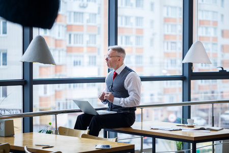 Adult male businessman, teacher, mentor working on a new project. Sits by a large window on the table. He looks at the laptop screen.の写真素材