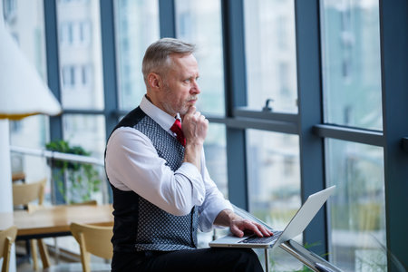 Adult male businessman, teacher, mentor working on a new project. Sits by a large window on the table. He looks at the laptop screen.の写真素材