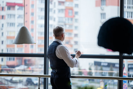 Smiling happy managing director thinks about his successful career development while standing with a cup of coffee in his hand in his office near the background of a window with copy spaceの写真素材