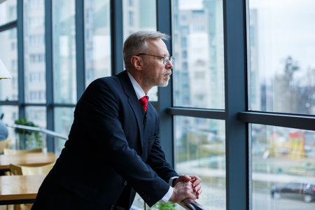 Adult male businessman, teacher, mentor looks out the large window of his office. Work in the office. Development of a new business projectの写真素材