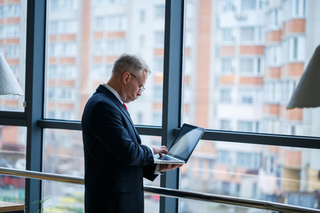 Smiling happy managing director thinks about his successful career development while standing with a laptop in his office near the background of a window with copy spaceの写真素材