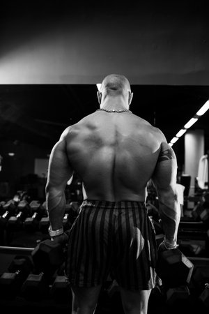 A strong, adult, fit, muscular male trainer poses for a photo shoot in a sports hall under the spotlights in sportswear, demonstrates his muscles and confidently holds dumbbells.の写真素材