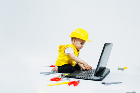 A two-year-old boy in a yellow vest on a white background sits with a laptop and plays with plastic...