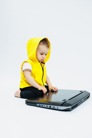 A cheerful 1-year-old boy in a yellow vest sits with a laptop on a white background. Modern...