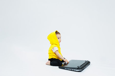 A cheerful 1-year-old boy in a yellow vest sits with a laptop on a white background. Modern...