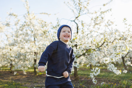 Cheerful boy 3 years old on a walk in a blooming garden. A little boy, 3 years old, in a sweater and a hat, runs through a blooming garden. Clothes for children aged 3 years. Emotions of child's joyの写真素材