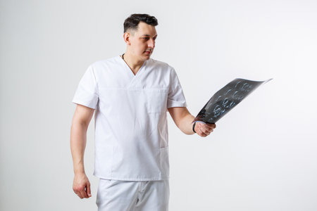 Young male doctor in white surgical suit holding and examining an MRI scan of the patient's head...