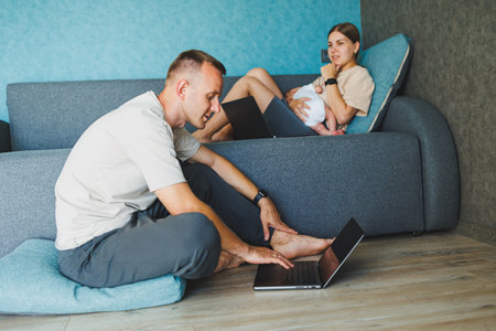 Parents with baby sitting on sofa at home with laptop and talking online. A young family with a child works on a laptop at home while sitting on the sofa.の写真素材