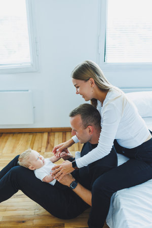 Happy family at home: father, mother and newborn son playing on white bed in bedroom. Young family...