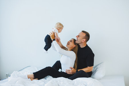 Happy family at home: father, mother and newborn son playing on white bed in bedroom. Young family...