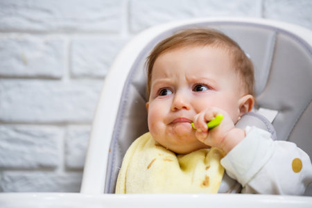 A nine month old smiling baby sits at a white table in a highchair and eats with a spoon from a bowl. Mom feeds the baby from a spoon. Blurred background. Healthy food for children. Children food.の写真素材