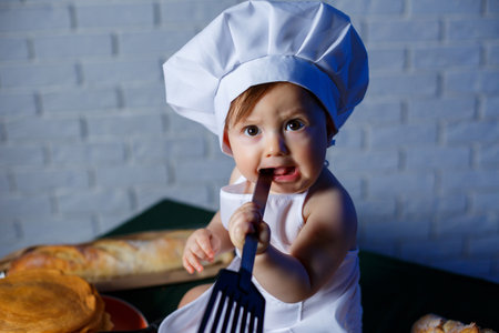 Little beautiful child dressed in an apron, children's cookery costume. Pancakes and fresh pastries on the tableの写真素材