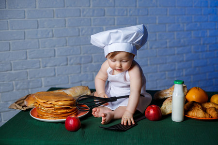 Little beautiful child dressed in an apron, children's cookery costume. Pancakes and fresh pastries on the tableの写真素材