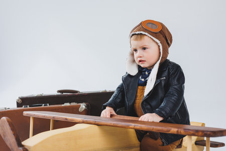 The boy is crying in a leather jacket and a pilot's hat, a wooden plane, brown suitcases. Children's wooden toys. Baby emotionsの写真素材