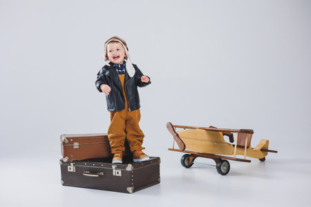 The boy is crying in a leather jacket and a pilot's hat, a wooden plane, brown suitcases. Children's wooden toys. Baby emotionsの写真素材