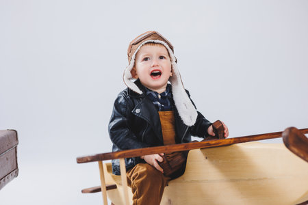 A happy boy in a helmet and a pilot's jacket stands near a wooden plane. Portrait of a child pilot, a child in a leather jacket. Wooden Toys. Eco plane from treeの写真素材
