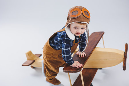 Little boy 3 years old plays with a wooden plane on a white background. In overalls and a pilot's...