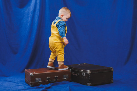 Ukrainian boy 3 years old in yellow overalls plays with suitcases. Modern child on a blue background. Happy child.の写真素材