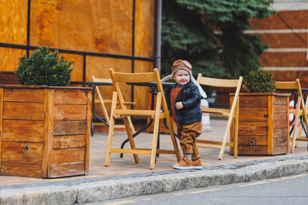 Stylish boy 3 years old in a leather jacket and brown trousers sits on the terrace near the cafe. Modern child. Children's fashion. Happy childの写真素材