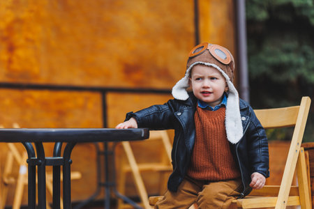 Stylish boy 3 years old in a leather jacket and brown trousers sits on the terrace near the cafe. Modern child. Children's fashion. Happy childの写真素材