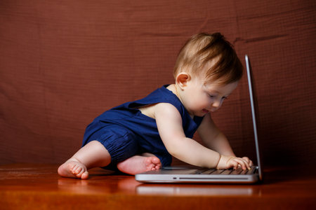 A small child 6-7 months old in a blue cotton overalls sits with an open laptop and watches educational gamesの写真素材