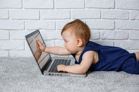 A small child 6-7 months old in a blue cotton overalls sits with an open laptop and watches educational gamesの写真素材