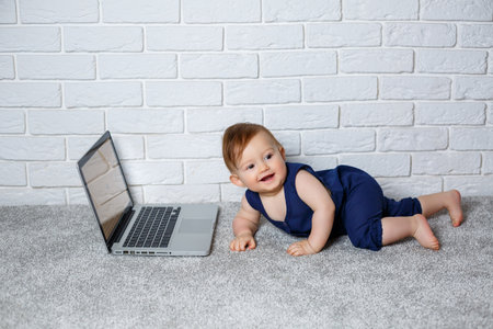 A small child 6-7 months old in a blue cotton overalls sits with an open laptop and watches educational gamesの写真素材