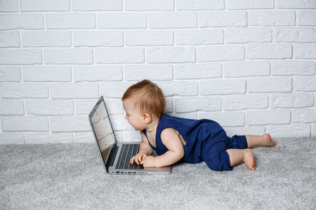 A small child 6-7 months old in a blue cotton overalls sits with an open laptop and watches educational gamesの写真素材