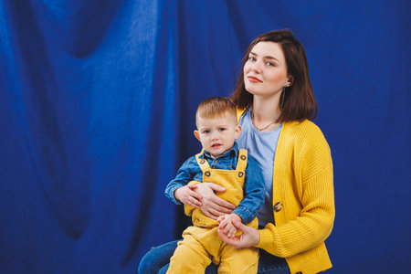 A young mother sits with a 3-year-old son in her arms, family relationships with a child. Children's education. Mom with son on a blue backgroundの写真素材