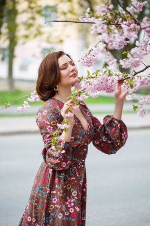 A woman walks along a spring street. The girl enjoys the scent of a blossoming tree. Beautiful woman in a dress with blooming cherry sakuraの写真素材