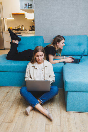 Two young female friends working at home sitting on the sofa at laptops. Beautiful girl students write reports on laptops.の写真素材