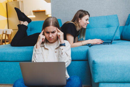 Two young female friends working at home sitting on the sofa at laptops. Beautiful girl students write reports on laptops.の写真素材