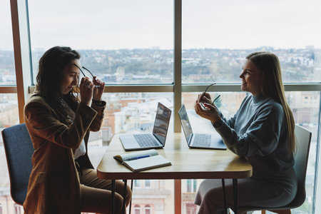 Two young female colleagues sitting with laptops near large windows, modern workspace. Female office workersの写真素材