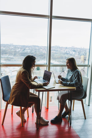 Two young female colleagues sitting with laptops near large windows, modern workspace. Female office workersの写真素材