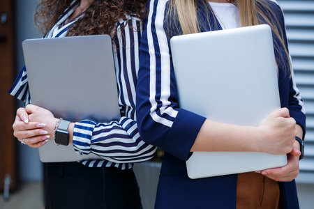 Portrait of two women businessmen who are standing with laptops in their hands, new project. Young business colleagues discuss office work. Successful female manager with glasses. Business conceptの写真素材