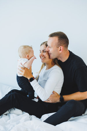 Happy family at home: father, mother and newborn son playing on white bed in bedroom. Young family...