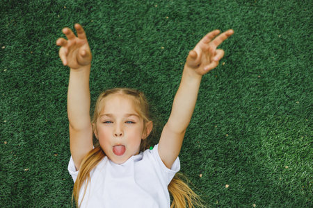 A little girl of 7-8 years old in a white T-shirt and sunglasses is sitting on the sports field. The...