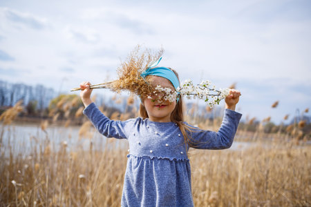 little girl child holds dry reeds and a branch with small white flowers in hands, sunny spring weather, smilling and joy of the childの写真素材