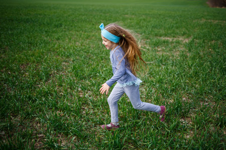 little girl child runs on green grass in the field, sunny spring weather, smile and joy of the child, blue sky with cloudsの写真素材