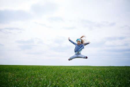 little girl child runs and jumps, green grass in the field, sunny spring weather, smile and joy of the child, blue sky with cloudsの写真素材