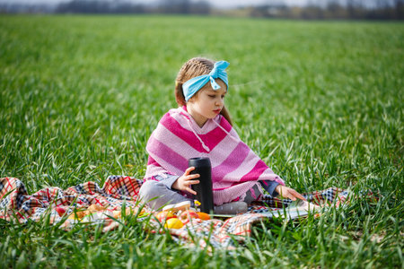 little girl child sits on a bedspread and reads a book with a fairy tale, green grass in the field, sunny spring weather, smile and joy of the child, blue sky with cloudsの写真素材