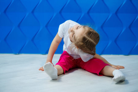 Little girl with pigtails in a white T-shirt on a blue background. She sits in a lotus position on the floor and smiles and waves her hands.の写真素材