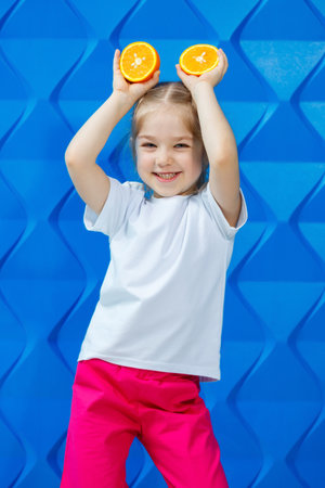 A little girl in a white T-shirt smiles and holds two halves of orange fruit in front of her eyes on an orange background. Concept: the fight against spring avitomnosis.の写真素材
