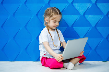 A modern child girl uses a laptop lying on the floor with legs looking at the camera. Funny kid on a bright blue wall background. Modern internet wireless technologyの写真素材