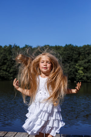 A six year old girl is wearing a white dress. Beautiful long hair. Walk near the river on the background of the forestの写真素材