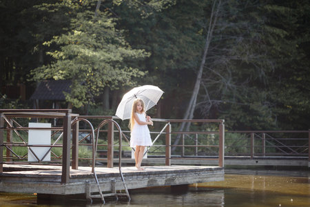 A beautiful girl of six years old in a short white dress stands on the masonry near the river and holds a white umbrella in her handsの写真素材