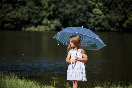 A little girl with long hair in a white dress stands on the bank of the river. Walk by the lake on a summer sunny dayの写真素材