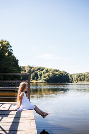 A 7-year-old girl with long blond hair by the lake sits on a clutch with legs in the water. She splashes her feet in the lake. Barefoot girl in a white dress with long hair.の写真素材