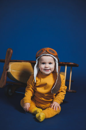 A little boy of 1 year in a yellow knitted suit and an aviator hat stands near a wooden helicopter. Children's wooden ecological toys made of antural woodの写真素材