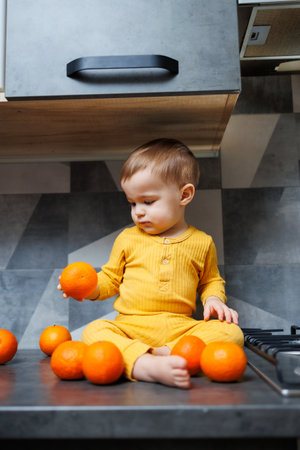 A little boy 1 year old in yellow clothes is sitting in the kitchen on the table with a plate of orange tangerines. Portrait of a cute one-year-old boy and sweet citrus fruits.の写真素材